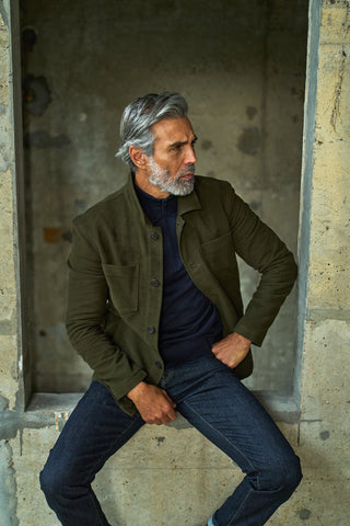 Man sitting in a rustic setting wearing an olive green jacket and dark jeans.