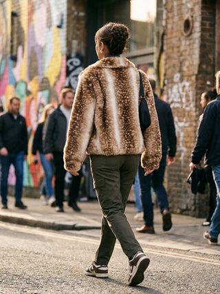 A person walking on a city street seen from behind, wearing a brown and beige faux-fur jacket, dark olive pants, and black-and-white sneakers. Other pedestrians and a graffiti-covered brick wall are visible in the background.