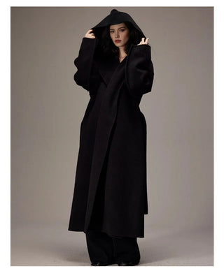A model wearing a long black double-breasted coat with a hood, paired with black trousers, set against a neutral gray studio background. Chinese text is displayed below the image.
