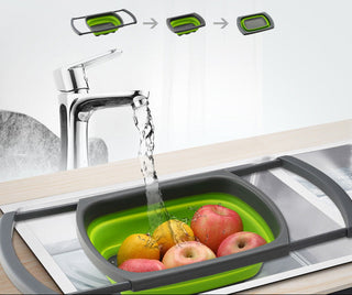 A green and gray collapsible fruit basket shown in a sink with water running from a faucet, holding apples and oranges. The image includes Chinese text and small diagram illustrations above showing the product's expandable and collapseable states.