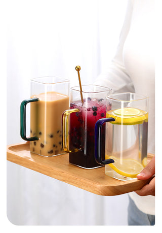 Three clear glass handled cups with colorful handles are arranged on a wooden tray, filled with iced tea, berry drink, and lemon water, held by a person in a white top against a soft white background.