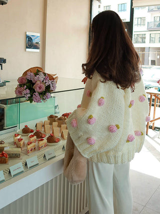 A woman viewed from behind wearing a cream knit cardigan with pink rose appliqués and white pants, standing in a bakery or cafe interior near a glass display case of pastries.