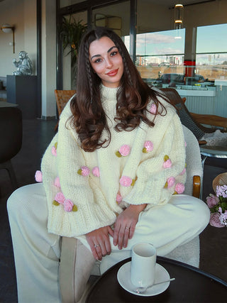 A woman sits indoors wearing a cream knit sweater with pink rose appliqués and white pants. A white cup and saucer sit on a small table in front of her, with a modern interior and large windows in the background.