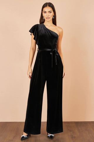 A woman modeling a black one-shoulder velvet jumpsuit with a matching waist tie, standing against a solid beige studio backdrop.
