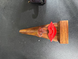 A small wooden figurine shaped like a female torso with a pointed brown body, a rectangular brown base, and glossy red lips, photographed on a dark gray surface.