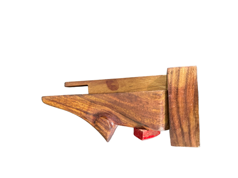 A handcrafted wooden object with a rich reddish-brown grain pattern, featuring a triangular front section, a vertical rectangular side piece, and a small red accent block, photographed on a dark textured surface.