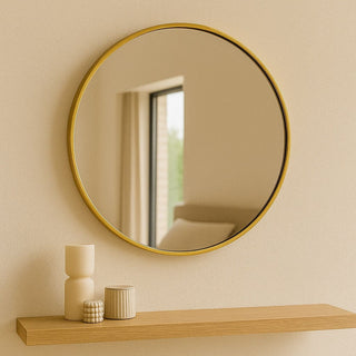 Round gold-framed mirror on a beige wall with a wooden shelf below.